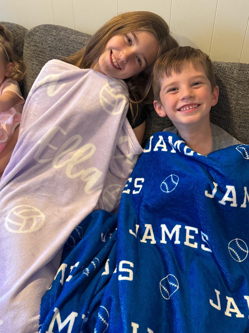 May include: Two children are sitting on a couch, each with a personalized fleece blanket. The girl's blanket is lavender with the name "Ella" and white volleyball designs. The boy's blanket is blue with the name "James" and white baseball designs.