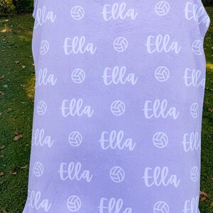 May include: A purple fleece blanket with the name "Ella" repeated throughout the design. White volleyball images are also repeated throughout the blanket.