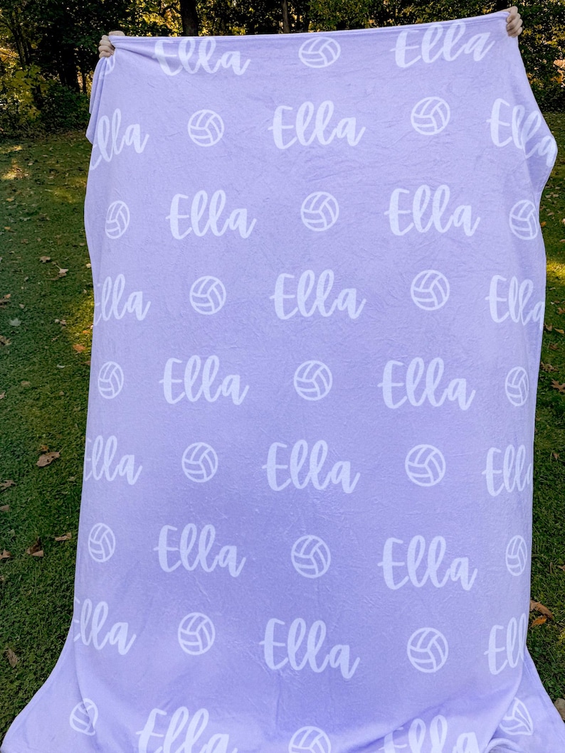 May include: A purple fleece blanket with the name "Ella" repeated throughout the design. White volleyball images are also repeated throughout the blanket.