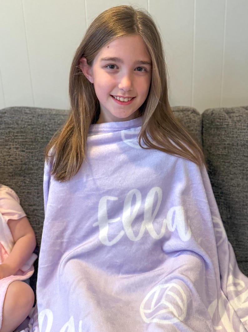 May include: A purple fleece blanket with the name "Ella" written in white cursive lettering. The blanket has a white volleyball design repeated throughout.