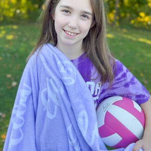 May include: A young person wearing a purple tie-dye shirt with the word "OH" on it, holding a pink and white volleyball. They are wrapped in a lavender fleece blanket with white lettering that says "Good Vibes Only".