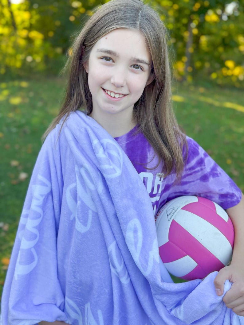 May include: A young person wearing a purple tie-dye shirt with the word "OH" on it, holding a pink and white volleyball. They are wrapped in a lavender fleece blanket with white lettering that says "Good Vibes Only".