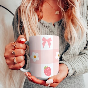 Könnte beinhalten: Eine weiße Keramiktasse mit einem rosa-weißen Karomuster. Die Tasse zeigt eine rosa Schleife, weiße Blumen und eine rote Erdbeere.
