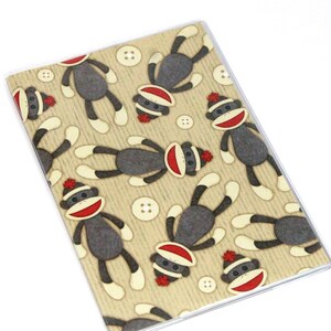 May include: A tan notebook cover with a repeating pattern of gray sock monkeys wearing red hats. The monkeys are in various poses and have white buttons on their bodies.