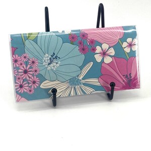 May include: A rectangular checkbook cover featuring a floral pattern with large light blue, pink, and white flowers on a teal background. The cover is held upright by a black metal display stand, showcasing the vibrant botanical print.