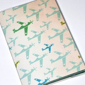 May include: A small notebook with a light blue and green airplane pattern on a cream background. The notebook has a clear plastic cover.