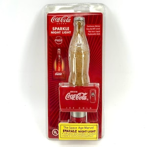 May include: Coca-Cola Sparkle Night Light in a clear plastic package. The night light is shaped like a Coca-Cola bottle and has a red base with the Coca-Cola logo. The packaging includes the text "Sparkle Night Light".