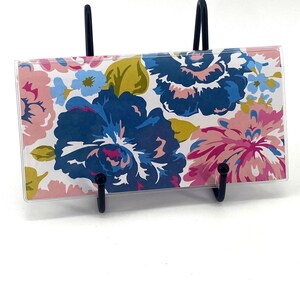 May include: A rectangular checkbook cover featuring a vibrant floral pattern with large dark blue, light pink, and yellow flowers on a white background, held upright by a black metal display stand.