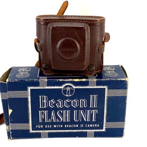 May include: A dark brown leather camera case with a circular embossed logo sits atop a rectangular blue cardboard box. The box features white text reading Beacon II FLASH UNIT FOR USE WITH BEACON II CAMERA and a lighthouse graphic icon.