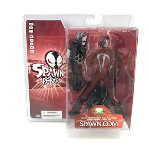 May include: A packaged action figure from the Spawn Reborn Series Two. The figure is in a clear plastic case with a red and white background. The figure is primarily red with white accents and includes a large gun and other accessories. The packaging includes the text "SPAWN.COM".