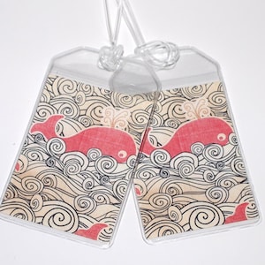 以下が含まれることがあります： ベージュの背景に黒い渦巻き模様が描かれた、赤いクジラのイラストが描かれた透明なプラスチック製の荷物タグが2つ。クジラは波の海を泳いでいます。