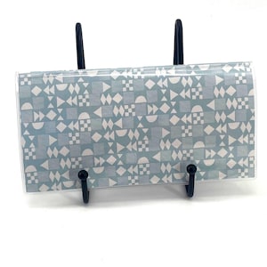 May include: A rectangular wallet featuring a light blue background with a repeating white geometric pattern of triangles, squares, diamonds, and semicircles. The wallet is held upright by a black metal display stand with two vertical supports.