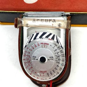 May include: A vintage Accura light meter featuring a circular silver dial with red and black numerical markings, including ASA and Polaroid settings. The device is encased in a brown leather protective sleeve with a clear plastic strap across the top.