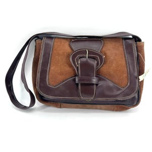 May include: A rectangular shoulder bag crafted from a combination of brown leather and tan suede. The bag features a front flap with a decorative leather buckle strap, contrast stitching, and a single shoulder strap attached to the sides.