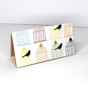 Puede incluir: Una tarjeta blanca con un patrón de jaulas de pájaros de colores. Las jaulas son de varios colores, incluyendo rojo, azul, verde y amarillo. Cada jaula tiene un pájaro negro dentro.