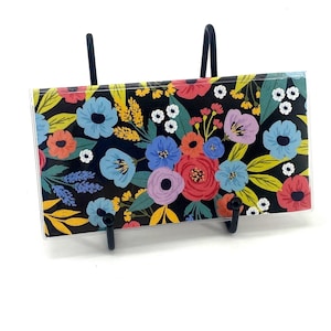 May include: A rectangular glass plaque featuring a vibrant floral pattern on a black background, displayed on a black metal stand. The design includes red, blue, purple, and yellow flowers with green leaves and small white blossoms.
