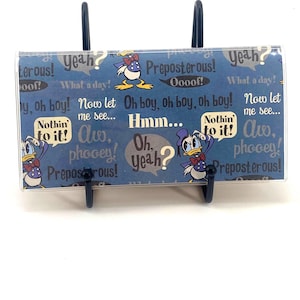 Op de afbeelding: Een blauw-witte Donald Duck-thema kaarthouder met een doorzichtige plastic voorkant. Het ontwerp bevat Donald Duck-illustraties en tekstballonnen met zinnen als "Oh boy!" en "Preposterous!" De houder wordt ondersteund door een zwarte metalen standaard.