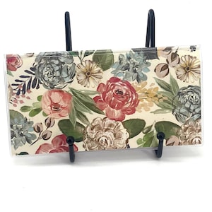 May include: A rectangular card featuring a floral pattern with red, pink, blue, and white flowers, green leaves, and dark stems. The card is encased in a clear plastic sleeve and is supported by a black metal display stand with two prongs.
