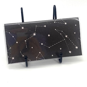 May include: A rectangular black card or envelope featuring a white celestial constellation pattern with various stars and connecting lines, held upright by a black metal wire display stand against a plain white background.