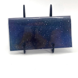 May include: A rectangular, clear acrylic photo holder with a dark blue and white galaxy design. The holder is supported by a black metal stand with two vertical posts and a curved base, suitable for displaying photos or artwork.