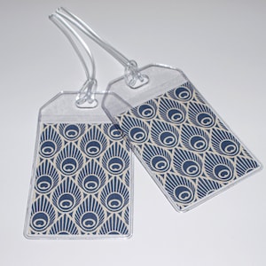 May include: Two clear plastic luggage tags with a blue and white peacock feather pattern. The tags have a string attached to them.
