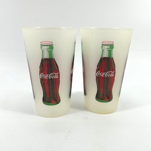 May include: Two frosted glass Coca-Cola glasses. Each glass features a red and green Coca-Cola bottle design. The glasses have a slightly tapered shape and are set against a white background.