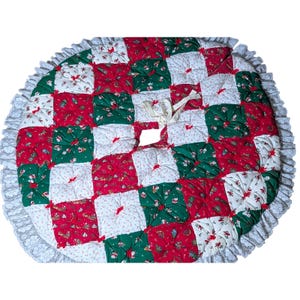 May include: A circular quilted Christmas tree skirt featuring a patchwork pattern of red, dark green, and white fabric squares with festive holiday prints. The skirt is finished with a white lace ruffle trim and includes white fabric ties.