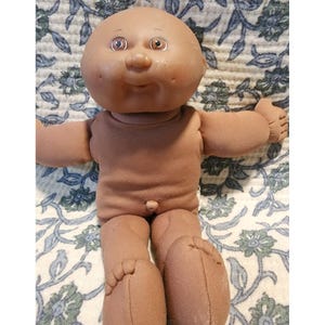 Pode incluir: Uma boneca de pele morena com uma cabeça grande e redonda, bochechas com covinhas e olhos castanhos. A boneca tem um corpo de tecido macio com um pequeno umbigo, braços estendidos e pernas para a frente, com dedos das mãos e pés costurados.