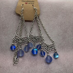 Op de afbeelding: Een paar zilveren oorhangers met blauwe, iriserende glazen kralen en een filigraanontwerp. De oorhangers bevinden zich op een kaart met de tekst "Handmade with love".