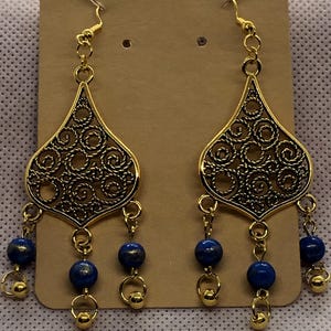 May include: Gold filigree dangle earrings with blue beads. The earrings feature a teardrop-shaped design with intricate details and three blue beads hanging from each earring.