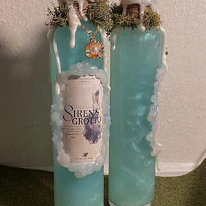 May include: Two glass bottles filled with a shimmering, light blue liquid. Each bottle has a cork stopper, adorned with moss and dripping white wax. One bottle has a label that reads "Siren's Grotto" and a gold seashell pendant.