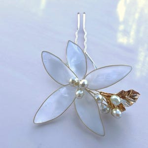 Puede incluir: Un pasador de pelo con forma de flor blanca con detalles de perlas y una hoja dorada. La flor tiene cinco pétalos con un borde dorado. Diseñado para ocasiones formales y bodas.