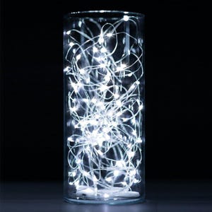 May include: A clear glass cylinder filled with white LED string lights. The lights are tightly packed and create a bright, glowing effect. The background is black, highlighting the illuminated lights. The overall theme is home decor and lighting.