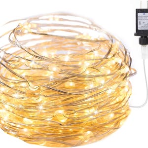May include: A bundle of warm white LED string lights with a silver wire. The lights are connected to a black power adapter with a white cord and a two-prong plug. The lights are tightly coiled, creating a dense, glowing effect.