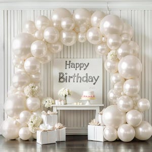 May include: A festive birthday scene with a large balloon arch in shades of white and cream. A white sign with "Happy Birthday" is centered, with a small cake and gifts. Hydrangeas and other floral arrangements add to the celebratory atmosphere.
