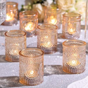 May include: A collection of amber-colored glass votive candle holders with intricate embossed patterns. Each holder contains a lit candle, casting a warm glow. The holders are arranged on a reflective surface, creating a romantic ambiance.