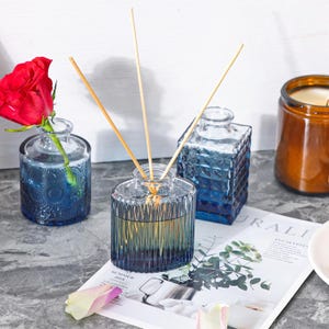 Puede incluir: Una variedad de botellas decorativas de vidrio en tonos azules y transparentes. Una botella contiene ca&ntilde;as de madera, otra una rosa roja. Tambi&eacute;n se ven una vela en un tarro marr&oacute;n y una revista con la palabra "Eucalyptus".