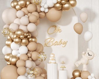 May include: A baby shower decoration featuring a balloon arch in shades of gold, white, and beige. The arch is adorned with clear balloons filled with gold confetti. The words "Oh Baby" are displayed in gold script, along with a teddy bear and a cake.