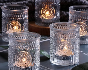 May include: A collection of clear glass votive candle holders. Each holder features an embossed floral design and contains a lit candle, creating a warm, inviting glow. The holders are arranged on a surface, showcasing their decorative details.