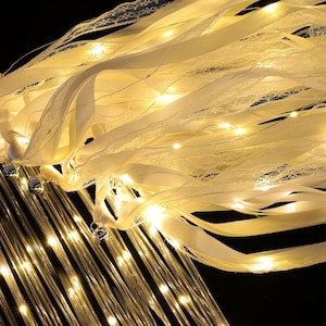 May include: Ivory-colored ribbons and lace with tiny, warm white LED lights. The ribbons are layered and flowing, with delicate lace detailing. Small silver bells are attached. The overall effect is soft and romantic, suitable for weddings or other celebrations.
