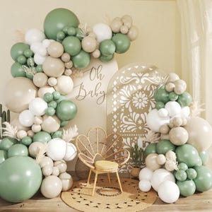 May include: A decorative baby shower scene with a balloon arch in shades of green, white, and beige. The words "Oh Baby" are written on a white sign. A woven chair and a round rug complete the setup.