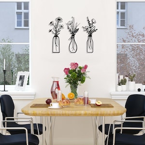 Puede incluir: Tres piezas de decoraci&oacute;n de pared de metal negro con arreglos florales estilizados en jarrones. Las piezas est&aacute;n montadas en una pared blanca sobre una mesa de comedor de madera con sillas negras.
