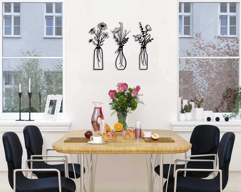 Puede incluir: Tres piezas de decoraci&oacute;n de pared de metal negro con arreglos florales estilizados en jarrones. Las piezas est&aacute;n montadas en una pared blanca sobre una mesa de comedor de madera con sillas negras.