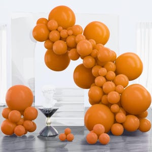 May include: Orange balloon arch and clusters of orange balloons. A white bust sits on a small table with a marble top and a black and gold base. The balloons are arranged in a decorative display.