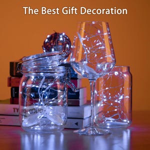 May include: Clear glass jars, a wine glass, and a round decorative sphere are illuminated with white fairy lights. The text "The Best Gift Decoration" is at the top. The jars and glass are on a table with books in the background.