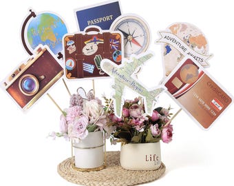 May include: A travel-themed centerpiece with paper cutouts of a suitcase, passport, camera, globe, compass, airplane, and credit card. The arrangement includes two small vases with pink and purple flowers, set on a woven mat.