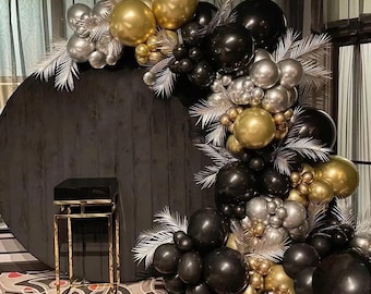 May include: A black, gold, and silver balloon arch with decorative white feathers. The arch is set against a dark circular backdrop with a gold and black side table. The balloons are in various sizes and metallic finishes.