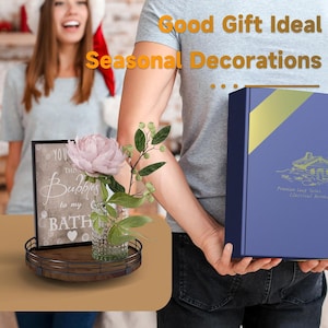 May include: A wooden tray with a black metal frame holds a clear glass jar with a pink flower and green leaves. A framed sign with the text "You add the bubbles to my bath" is on the tray. A person is holding a blue box with gold trim and a gold logo.