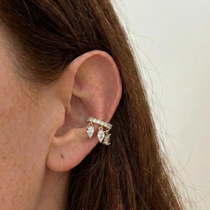 May include: Gold-toned ear cuff with sparkling clear crystals. The cuff features a row of small crystals and three dangling charms: two pear-shaped and one rectangular. The jewelry is worn on the ear.