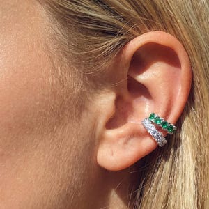 May include: Ear cuff earrings with a row of sparkling white stones and a row of green stones. The earrings are worn on the ear. The skin is fair and the hair is blonde.